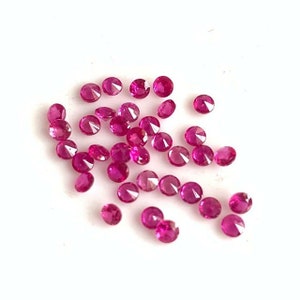 May include: A collection of round, faceted ruby gemstones. The rubies are a vibrant pink colour and are scattered on a white surface.