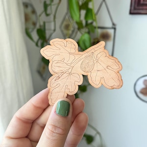 May include: A wooden fig leaf and fruit cut-out. The light brown wood carving depicts a fig with detailed leaf veins and fruit texture. The fig branch is held by a person with green nail polish. The background includes plants and decorative frames.