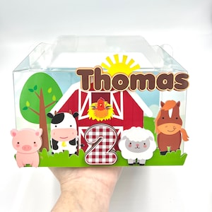 Puede incluir: Una caja de plástico transparente con un diseño de granja. La caja presenta un granero rojo, animales de dibujos animados y el nombre "Thomas" con el número "2". El diseño incluye un cerdo, una vaca, una oveja y un caballo.