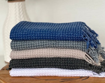 Könnte beinhalten: Ein Stapel von sechs Waffelhandtüchern in verschiedenen Farben: Marineblau, Grau, Beige, Anthrazit und Weiß. Die Handtücher haben eine strukturierte Oberfläche und Fransen. Die Handtücher sind ordentlich auf einer Holzoberfläche gestapelt.