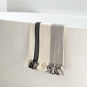 Puede incluir: Una toalla de ba&ntilde;o blanca con rayas negras y borlas colgando del borde de una ba&ntilde;era blanca.