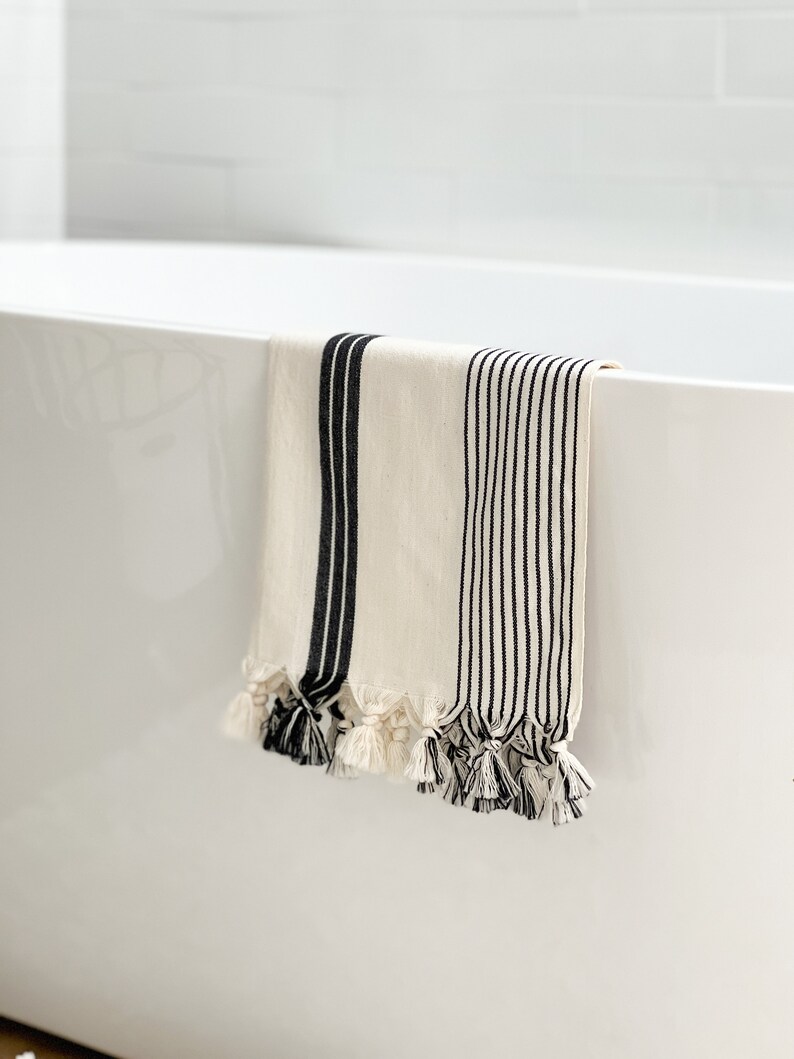 Puede incluir: Una toalla de ba&ntilde;o blanca con rayas negras y borlas colgando del borde de una ba&ntilde;era blanca.