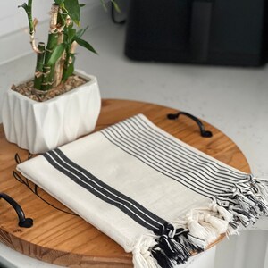 Puede incluir: Una toalla de algod&oacute;n a rayas blancas y negras con flecos en una bandeja de madera. La toalla est&aacute; doblada y tiene un patr&oacute;n de rayas blancas y negras.