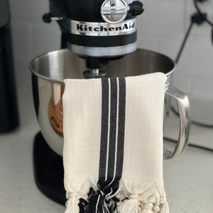 Puede incluir: Una batidora de pie KitchenAid negra con un taz&oacute;n plateado y una toalla de cocina blanca con rayas negras y borlas colgando del lado.