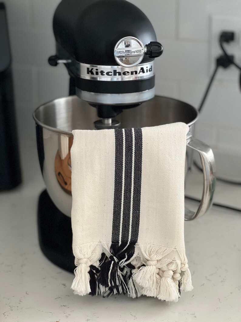 Puede incluir: Una batidora de pie KitchenAid negra con un taz&oacute;n plateado y una toalla de cocina blanca con rayas negras y borlas colgando del lado.