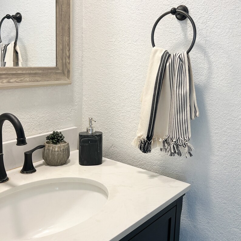 Puede incluir: Un toallero de metal negro con dos toallas rayadas blancas y negras colgadas. El toallero est&aacute; montado en una pared blanca en un ba&ntilde;o. Un mostrador blanco con un lavabo negro y un dispensador de jab&oacute;n negro est&aacute; en primer plano.