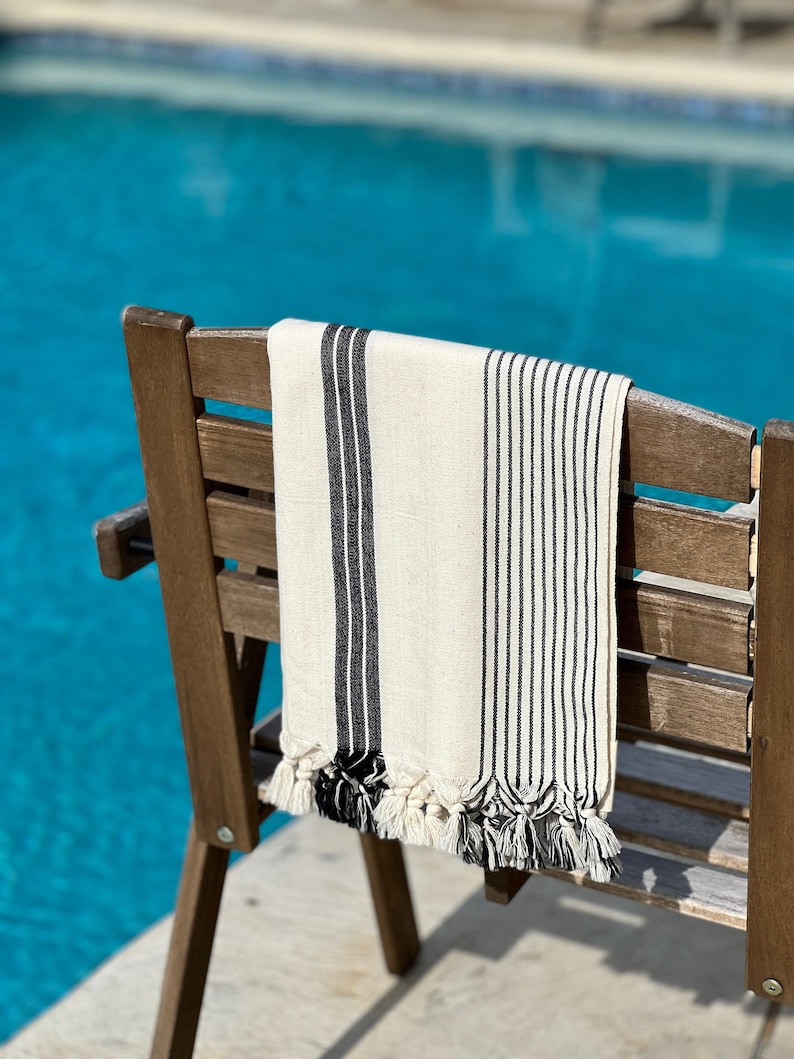 Puede incluir: Una toalla de playa a rayas blancas y negras con borlas colgando de una silla de madera junto a una piscina.