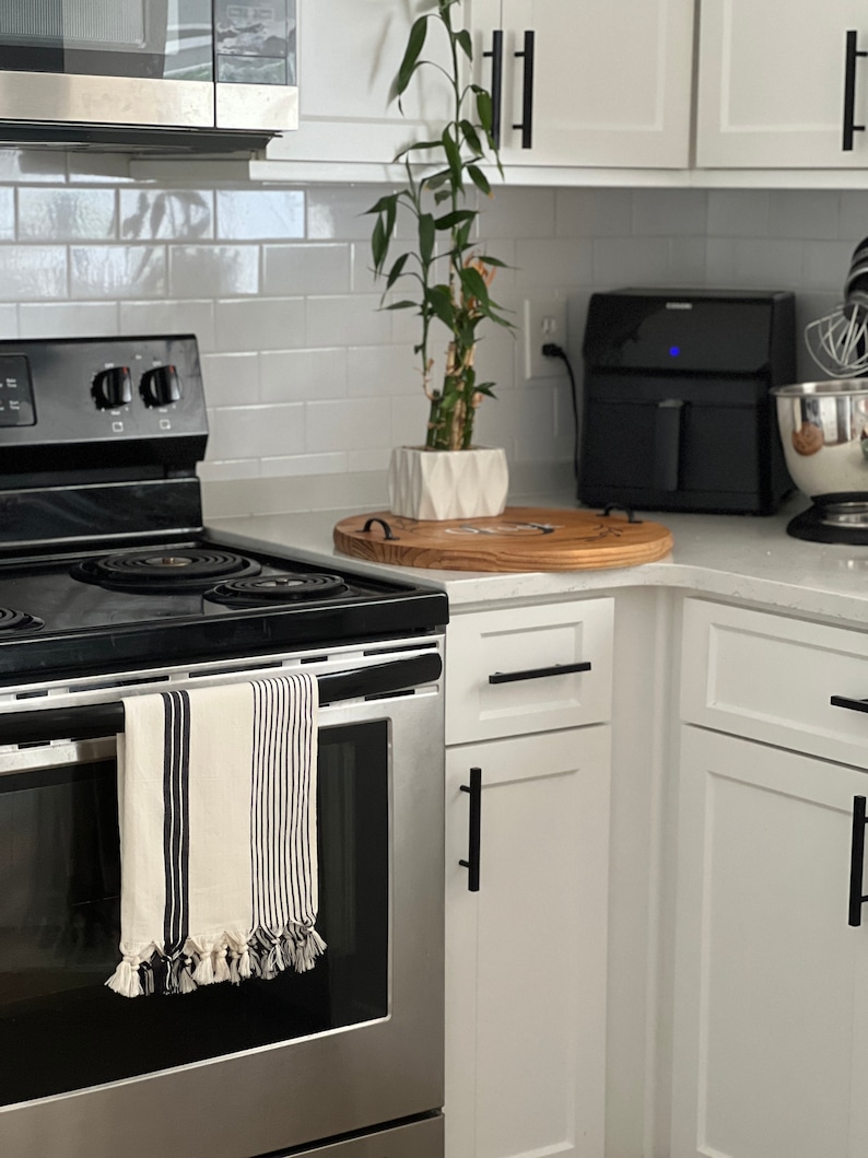 Puede incluir: Un horno de acero inoxidable con una toalla de cocina a rayas blancas y negras colgada del asa. El horno est&aacute; en una cocina moderna con gabinetes blancos y herrajes negros.