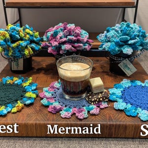 May include: Three crocheted flower arrangements in pots, with names "Forest", "Mermaid", and "Sky" written on the surface below.  The flowers are in shades of green, blue, and purple.  A cup of coffee sits on a crocheted coaster in the center.