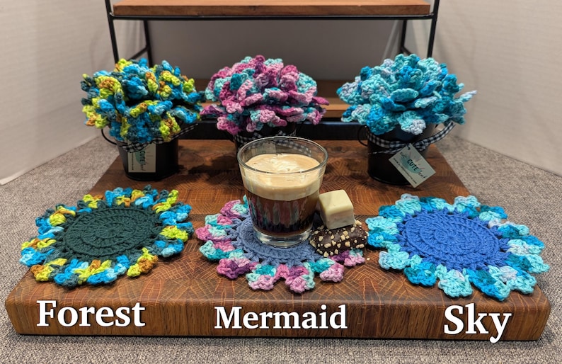 May include: Three crocheted flower arrangements in pots, with names "Forest", "Mermaid", and "Sky" written on the surface below.  The flowers are in shades of green, blue, and purple.  A cup of coffee sits on a crocheted coaster in the center.
