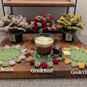 May include: Three crocheted plant pot covers with different colored flowers. The covers are pink and green, green and red, and green and yellow. The covers are sitting on a wooden surface with a cup of coffee and a few chocolate candies.
