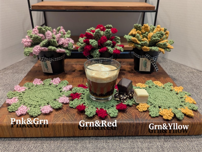 May include: Three crocheted plant pot covers with different colored flowers. The covers are pink and green, green and red, and green and yellow. The covers are sitting on a wooden surface with a cup of coffee and a few chocolate candies.