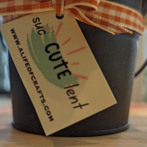 May include: A small black metal bucket with a white and orange checkered ribbon and a tag that says "Suc Cute Lent" and "www.alifeofcrafts.com".