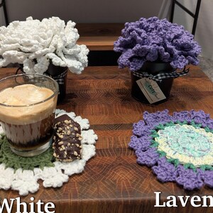 May include: Two crocheted flower arrangements in pots, one white and one lavender, with a crocheted coaster and a cup of coffee on a wooden surface. The lavender flower arrangement has a tag that says "Cute Craft".