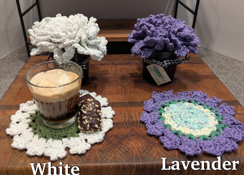 May include: Two crocheted flower arrangements in pots, one white and one lavender, with a crocheted coaster and a cup of coffee on a wooden surface. The lavender flower arrangement has a tag that says "Cute Craft".