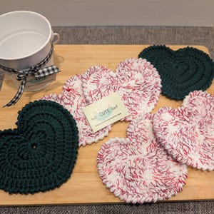 May include: Five crocheted heart-shaped coasters. Two are green, and three are white with pink stripes. The coasters are arranged on a wooden surface. The text "The Crafty Nest" is visible on a white tag.