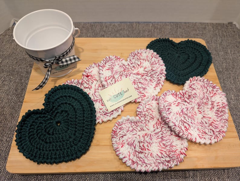 May include: Five crocheted heart-shaped coasters. Two are green, and three are white with pink stripes. The coasters are arranged on a wooden surface. The text "The Crafty Nest" is visible on a white tag.