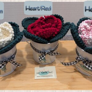 May include: Three crocheted flower arrangements in white, red, and pink, each in a white metal pot with a black and white checkered ribbon. The pots are on a wooden surface. The flowers are labeled "Heart/Cream", "Heart/Red", and "Heart/Mix".