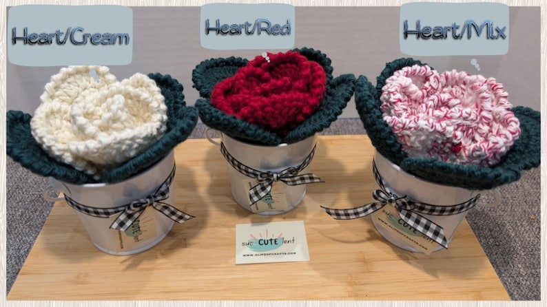 May include: Three crocheted flower arrangements in white, red, and pink, each in a white metal pot with a black and white checkered ribbon. The pots are on a wooden surface. The flowers are labeled "Heart/Cream", "Heart/Red", and "Heart/Mix".