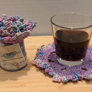 May include: A purple crocheted coaster with a glass of dark liquid on top. The coaster has a multi-colored design with pink, blue, and green accents. The glass is clear and has a rounded shape. The coaster is sitting on a wooden surface. The text on the tag reads "suc CUTE lent www.ALIFEofCRAFTS.com".