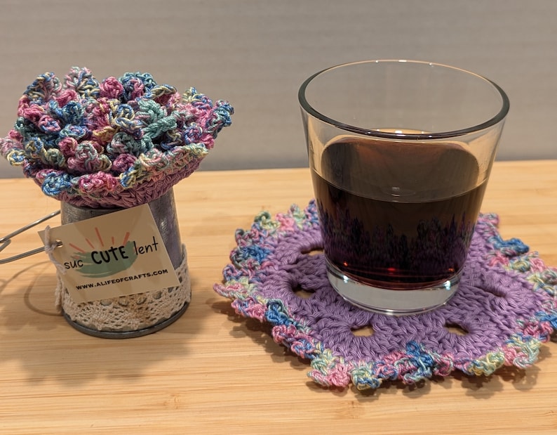 May include: A purple crocheted coaster with a glass of dark liquid on top. The coaster has a multi-colored design with pink, blue, and green accents. The glass is clear and has a rounded shape. The coaster is sitting on a wooden surface. The text on the tag reads "suc CUTE lent www.ALIFEofCRAFTS.com".