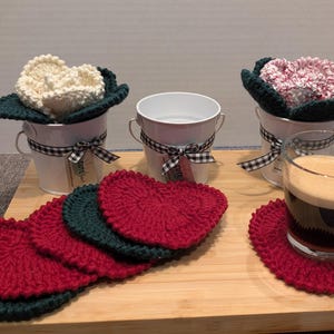May include: Five crocheted red and green coasters with a white, pink, and cream crocheted flower in a white metal bucket on a wooden surface. A glass of dark liquid sits on a red coaster.
