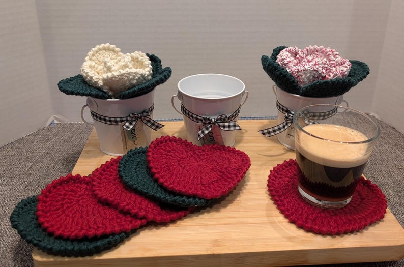 May include: Five crocheted red and green coasters with a white, pink, and cream crocheted flower in a white metal bucket on a wooden surface. A glass of dark liquid sits on a red coaster.