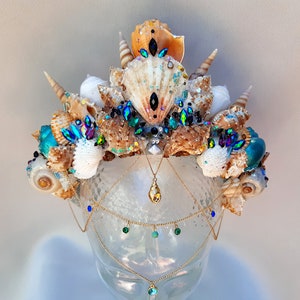 Può includere: Una corona di conchiglie artigianale a tema sirena. La corona presenta varie conchiglie, gemme iridescenti blu e verdi e dettagli di catene color oro. La corona è esposta su una testa di manichino trasparente.