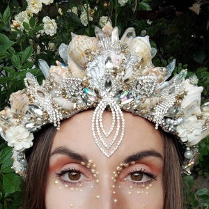 Puede incluir: Una corona artesanal con temática de sirena que presenta conchas marinas, cristales y brillantes pedrería. La corona tiene una combinación de colores plateado y dorado, con detalles florales blancos. El diseño es intrincado y detallado.