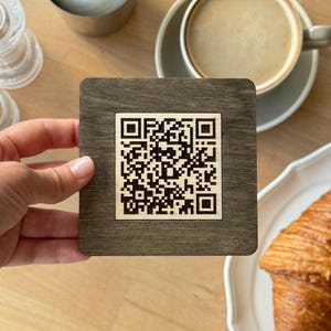 Könnte beinhalten: Ein quadratischer Holzuntersetzer mit einem QR-Code wird über einem Holztisch gehalten. Eine Tasse Kaffee, ein Croissant auf einem weißen Teller und Besteck befinden sich ebenfalls auf dem Tisch. Der Untersetzer ist braun mit hellem Rand.