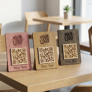 Könnte beinhalten: Drei hölzerne QR-Code-Ständer in Rosa, Beige und Braun, jeweils mit einem QR-Code und dem Text "Your Logo" und "Your Text". Die Ständer stehen auf einem Holztisch in einem Café.