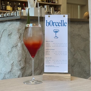Puede incluir: Un cóctel en una copa alta con un degradado rojo y transparente, sobre una mesa. Un menú con el nombre "bOrcelle" está en el fondo. La barra tiene estantes con botellas y adornos decorativos de pájaros.