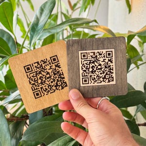 Puede incluir: Dos posavasos cuadrados de madera, uno marrón claro y otro gris oscuro, cada uno con un código QR. Los posavasos se sostienen frente a una planta verde. Los códigos QR son en blanco y negro.