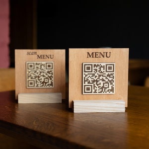Könnte beinhalten: Zwei hölzerne Menühalter mit QR-Codes. Einer trägt die Aufschrift "scan MENU" und der andere "MENU". Die Halter sind aus hellbraunem Holz gefertigt und stehen auf kleinen Holzständern. Der Hintergrund ist eine verschwommene Mischung aus Rosa und Schwarz.