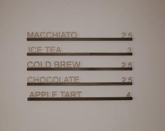 May include: A wooden sign with a menu of drinks and prices. The menu includes Macchiato, Ice Tea, Cold Brew, Chocolate, and Apple Tart. The prices are 2.5, 3, 2.5, 2.5, and 4.