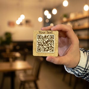 May include: A square wooden coaster with a QR code and the text "Your Text" engraved on it. The coaster is held in a hand against a blurred background. The coaster is approximately 3 inches by 3 inches.