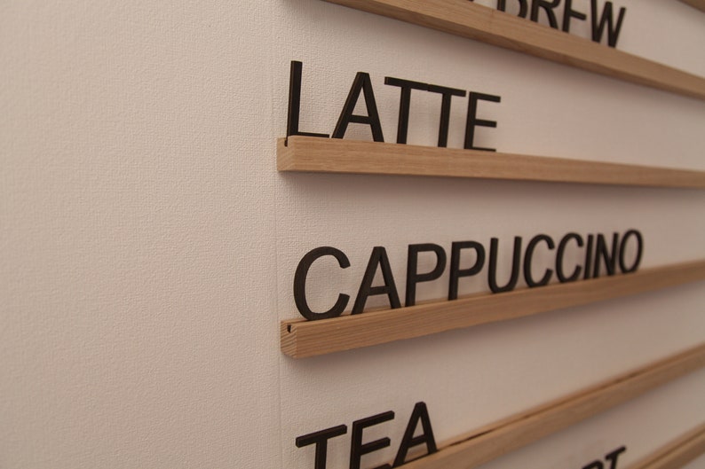 K&ouml;nnte beinhalten: Ein Holzschild mit schwarzen Buchstaben, die die W&ouml;rter "Brew", "Latte", "Cappuccino" und "Tea" buchstabieren.