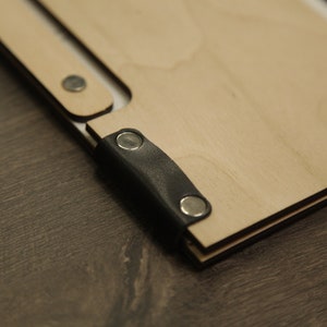 May include: A wooden notebook with a black leather strap and metal clasps. The notebook is closed and lying on a wooden surface.