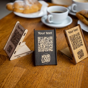 Può includere: Tre supporti in legno con codici QR e il testo "Your text" e "Your logo" inciso su di essi. I supporti sono su un tavolo di legno.
