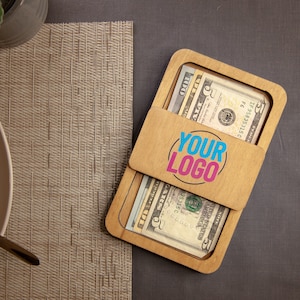 May include: A wooden money holder with a sliding lid, containing several U.S. dollar bills. The lid features the text "YOUR LOGO" in a colorful design. The holder is on a dark surface.