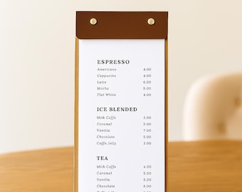 May include: A wooden menu holder with a brown leather top, displaying a coffee and tea menu. The menu lists espresso, ice blended, and tea options with prices. The holder is on a wooden table.