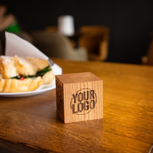 Può includere: Un cubo di legno con la scritta "YOUR LOGO" incisa su un lato è appoggiato su un tavolo di legno. Un panino al croissant su un piatto bianco è sullo sfondo. Il cubo è di colore legno naturale.