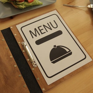 Puede incluir: Una cubierta de men&uacute; de madera con un inserto de men&uacute; en blanco y negro. El inserto de men&uacute; tiene la palabra "MENU" en texto negro y una ilustraci&oacute;n en blanco y negro de una campana.