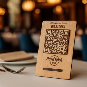 Puede incluir: Un portamenús de madera con un código QR y el logotipo de Hard Rock Cafe. El soporte tiene las palabras "SCAN for our Online MENU" en la parte superior. El fondo es un restaurante con mesas y sillas borrosas.