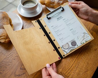 Puede incluir: Un portamenús de madera con una banda elástica negra y clips metálicos. El menú está impreso con la palabra "MENÚ" y una lista de artículos de comida y bebida.