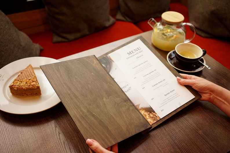 May include: A wooden menu cover with a white menu inside. The menu has the words "MENU" and "restaurant bar" printed on it. The menu is open to a page with a list of items and prices. A person is holding the menu with their right hand.