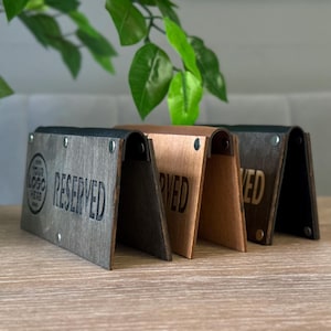 Puede incluir: Tarjetas de mesa de madera en varios tonos de marrón y gris, con la palabra "RESERVED" grabada en la parte delantera. Las tarjetas tienen una parte superior curvada y están sujetas con pequeños remaches metálicos.