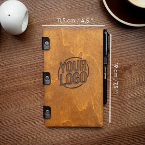 Könnte beinhalten: Ein Holz-Menüdeckel mit schwarzen Metallverschlüssen und dem eingravierten Text "YOUR LOGO" in einem Kreis. Der Deckel misst 19 cm x 11,5 cm.
