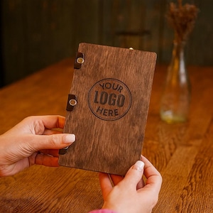 May include: A dark wooden menu cover with the text "YOUR LOGO HERE" in a circular design. The cover is held by hands and has leather straps with metal rivets. The wood has a rich, brown colour.