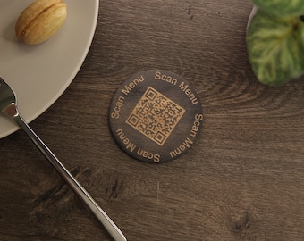 Op de afbeelding: Een ronde houten onderzetter met een bruine afwerking. De onderzetter heeft een QR-code in het midden en de tekst "Scan Menu" die herhaald wordt rond de rand.